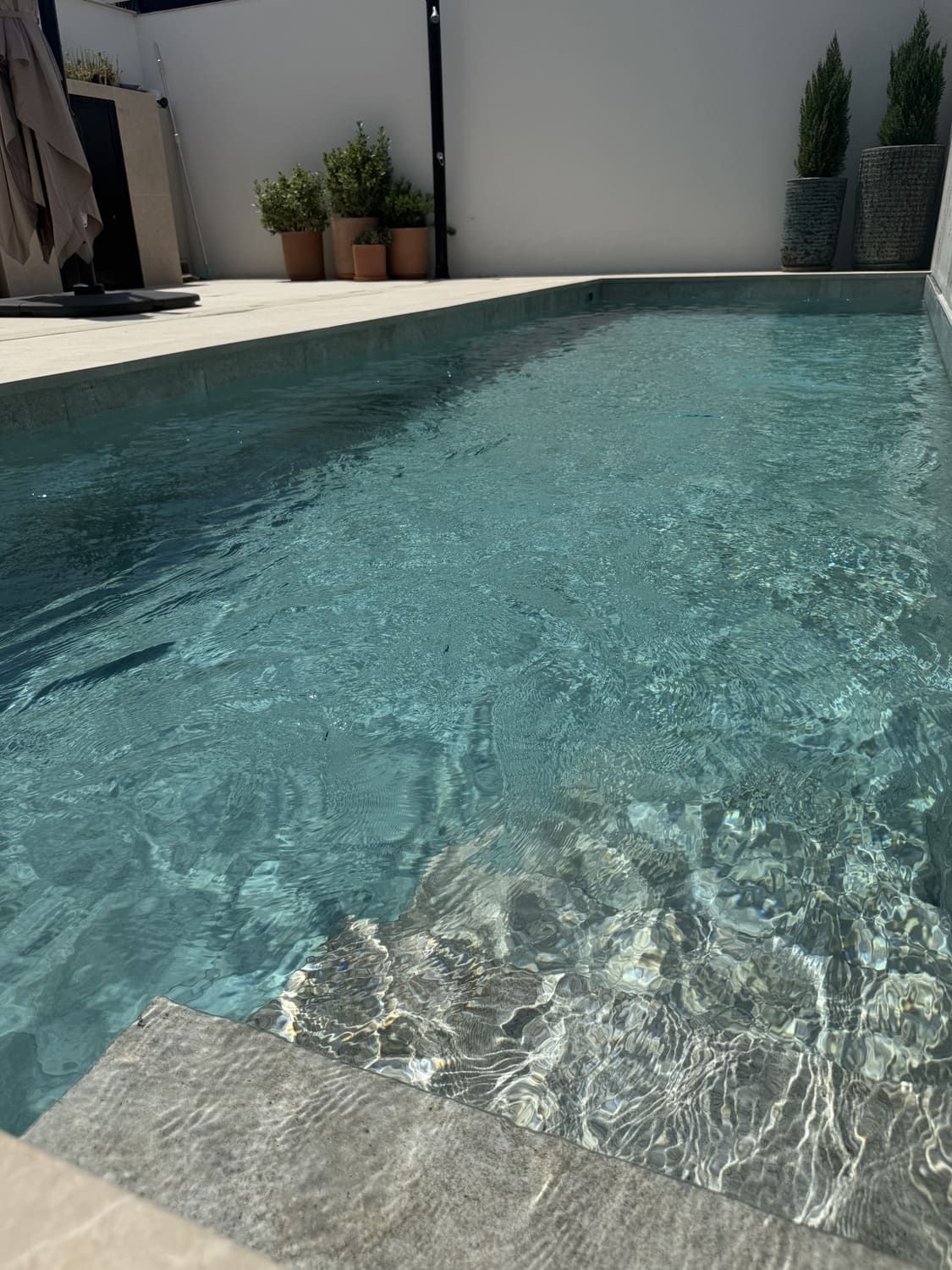 Piscina de agua azul cristalina y escalones de piedra, rodeada de una pared blanca y plantas en macetas al sol.