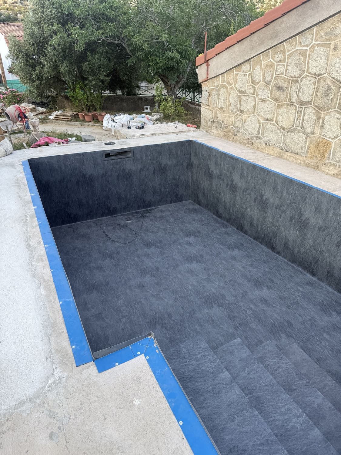 Piscina rectangular vacía, baldosas grises, cinta azul en los bordes, fondo de pared de piedra.