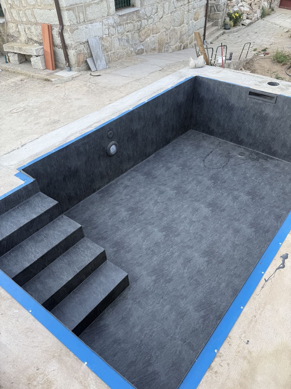 Piscina vacía rectangular con interior gris oscuro, escalones y borde azul, en un entorno de hormigón.