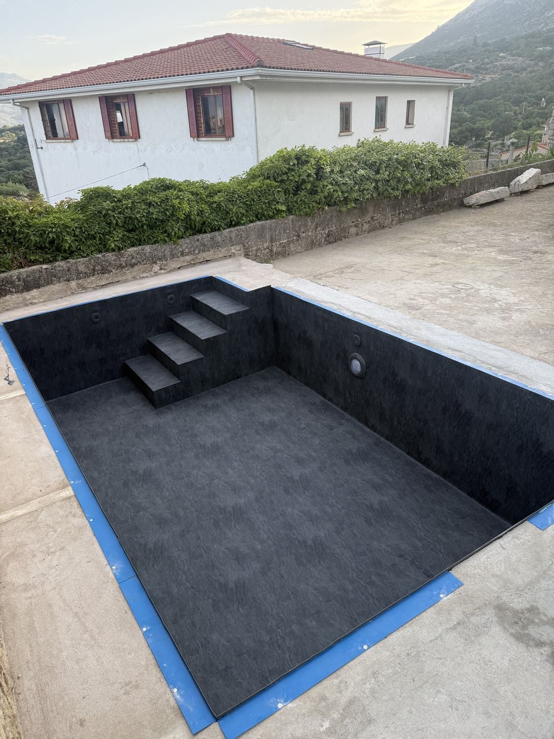 Piscina vacía con interior oscuro y escalones, delimitada por cinta azul. Al fondo se ve una casa blanca.