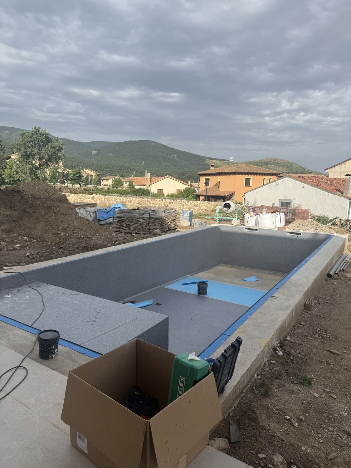 Sitio de construcción con una piscina rectangular enterrada; paredes grises, sellador azul; edificios, colinas y cielo nublado en el fondo.