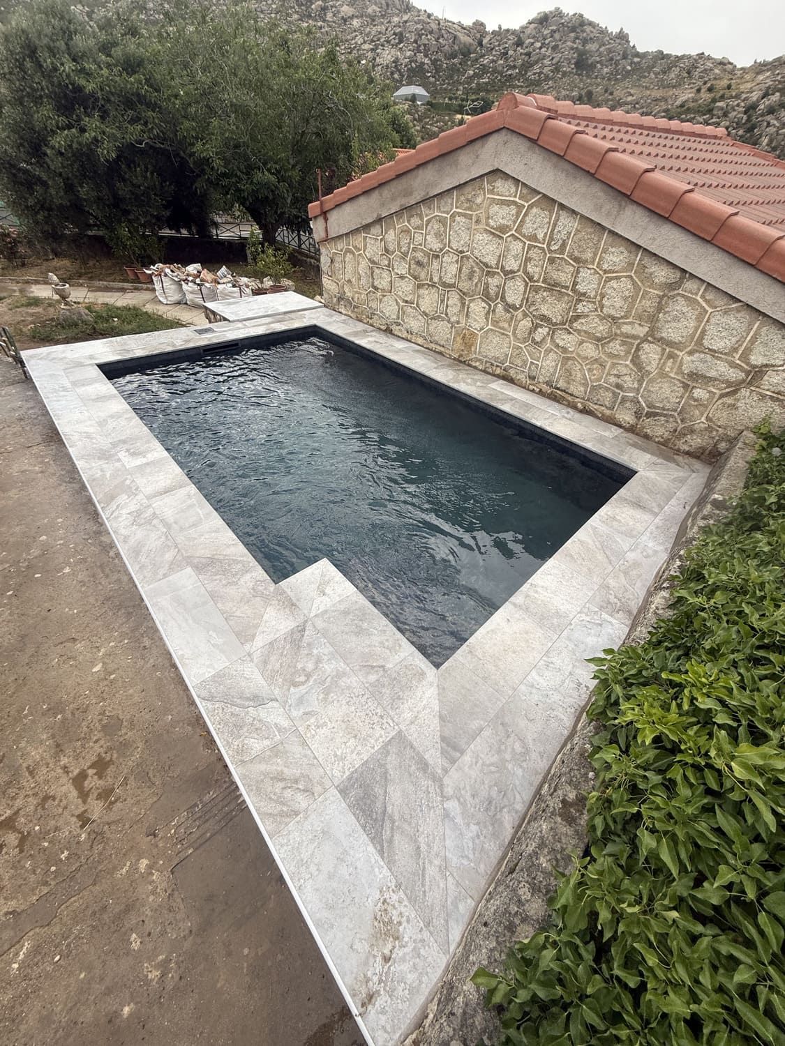 Pequeña piscina rectangular con escalones de piedra y moldura, junto a un muro de piedra, bajo techo de tejas rojas.