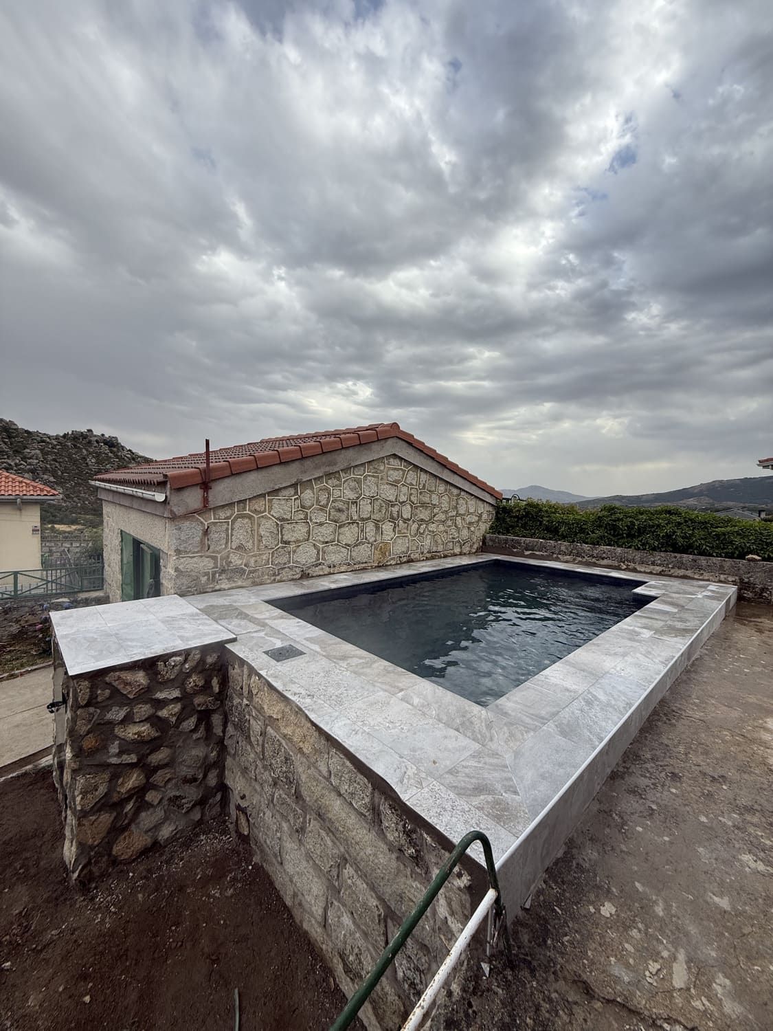 Piscina rectangular con borde de piedra, junto a una construcción de piedra, bajo cielo nublado.