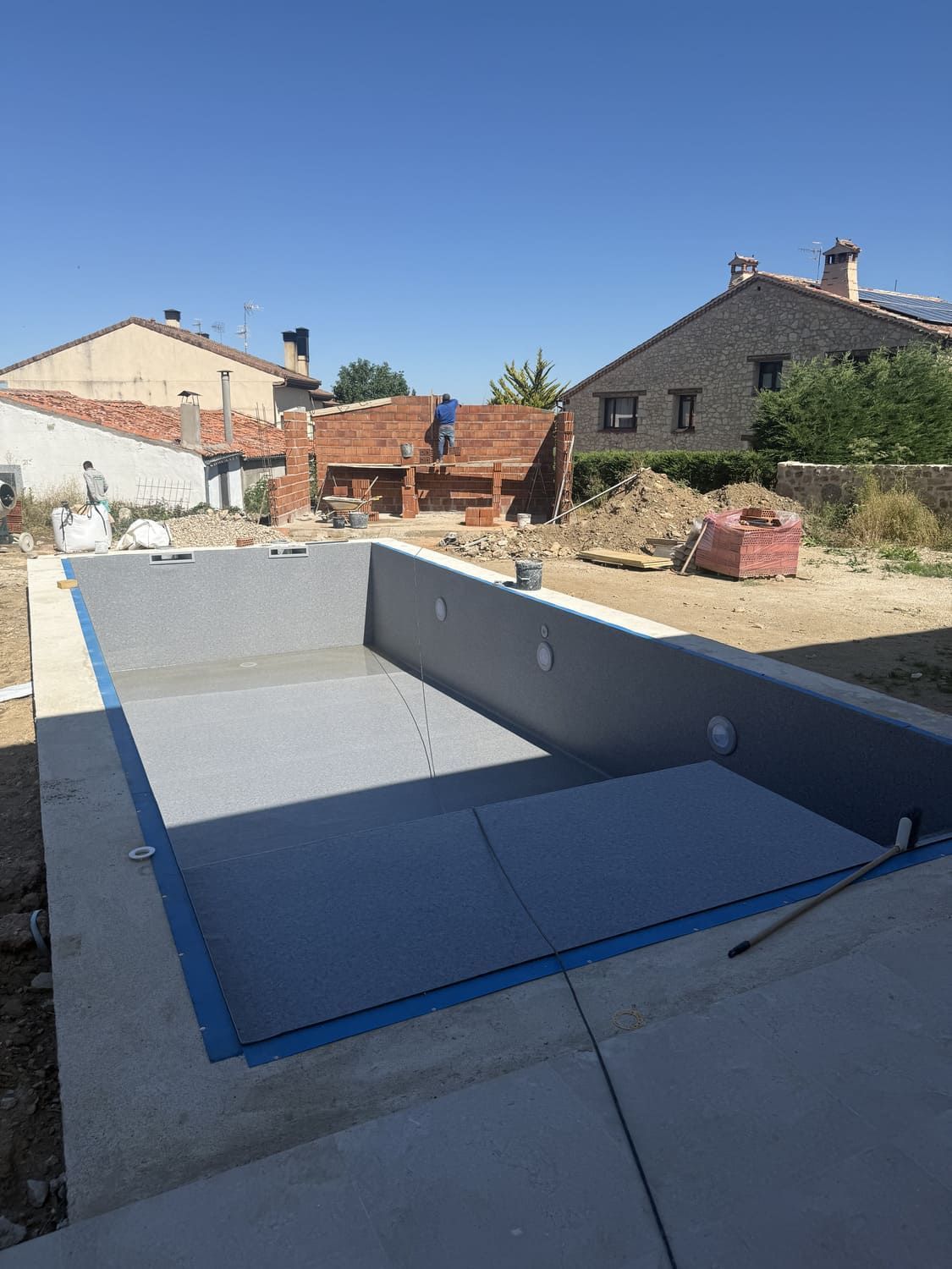 Una piscina en construcción con paredes interiores grises y una estructura de ladrillo al fondo bajo un cielo azul.