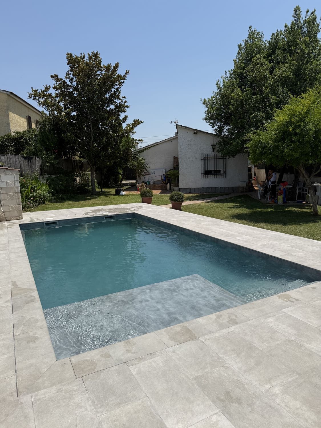 Piscina rectangular de agua azul y patio de piedra, rodeada de árboles y un patio trasero soleado.