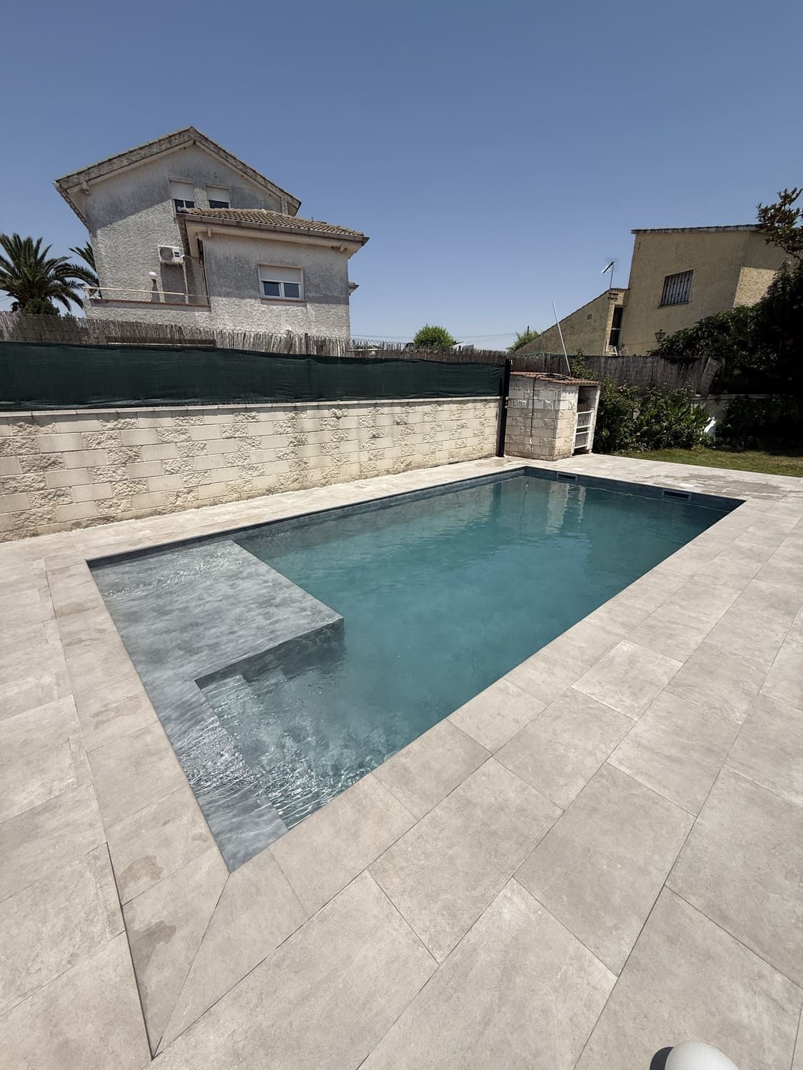 Piscina rectangular con escalones de piedra gris y patio circundante. Casas y cielo azul al fondo.