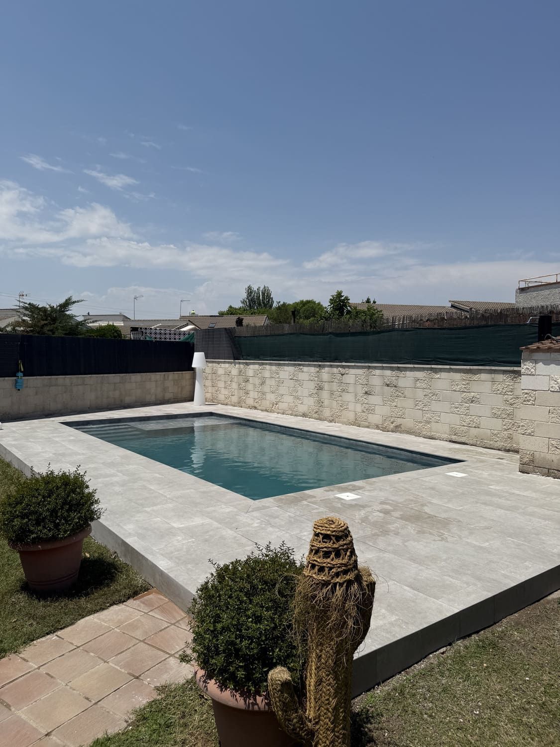 Piscina con patio de piedra, rodeada de césped, plantas en macetas y un muro de piedra bajo un cielo azul.