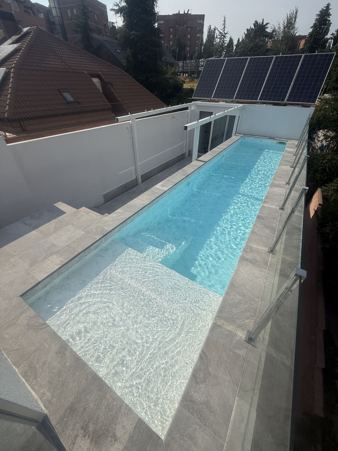 Piscina larga y rectangular con agua azul claro y escalones de baldosas blancas. Está rodeada por una pared blanca con paneles solares.