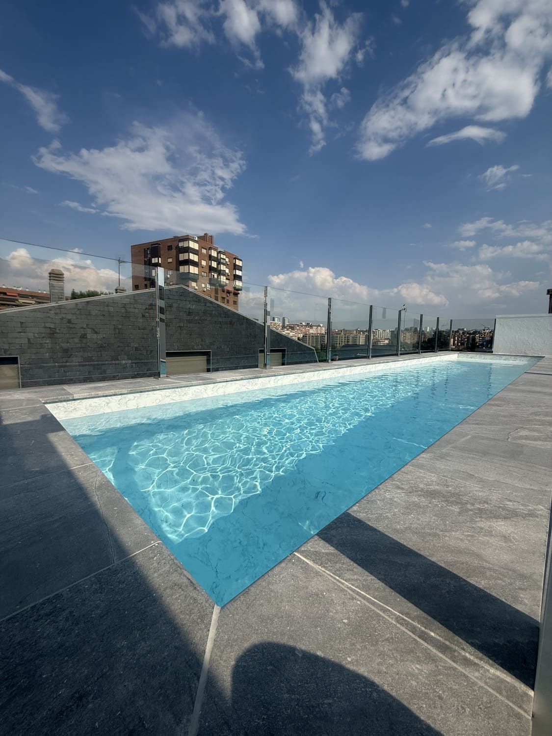 Piscina en la azotea con agua azul cristalina y cielo soleado. Marco de piedra gris y edificio al fondo.
