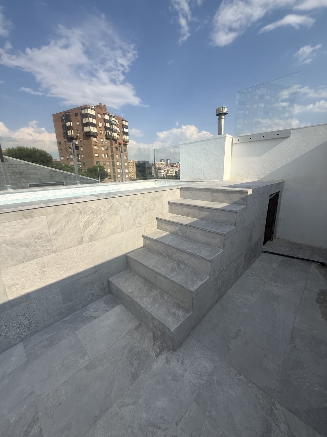 Azotea exterior con escalones de baldosas grises, paredes blancas y un edificio de varios pisos al fondo.