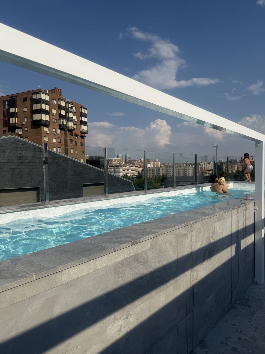 Piscina en la azotea con gente nadando, con vistas al horizonte de la ciudad bajo un cielo azul con nubes blancas.