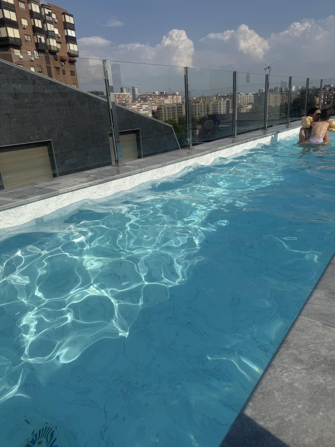 Piscina en la azotea con agua cristalina, vista a la ciudad, cielo azul y una persona nadando.