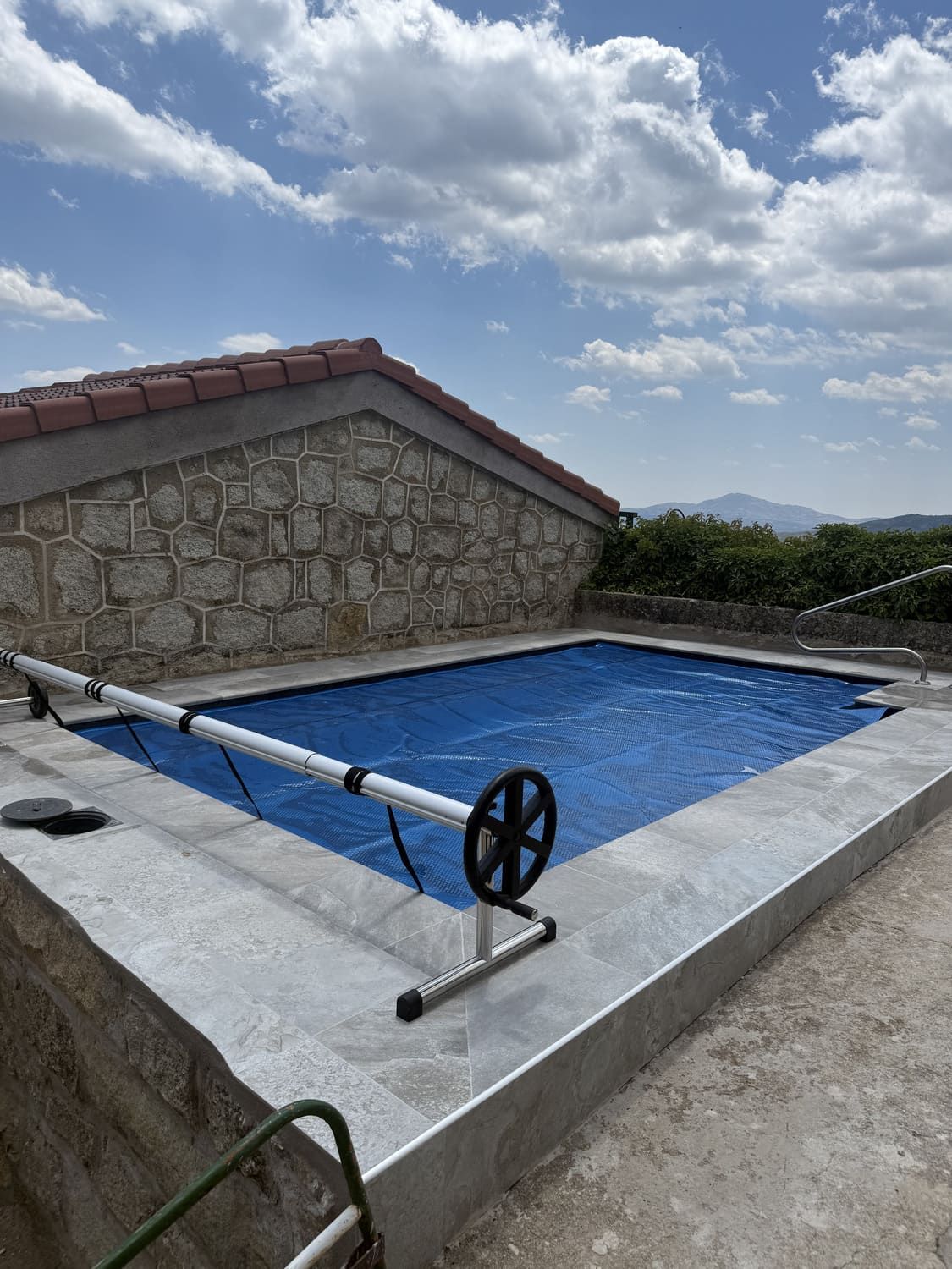 Piscina rectangular con cubierta azul en un patio de hormigón, con muro y techo de piedra al fondo. Cielo brillante con nubes.