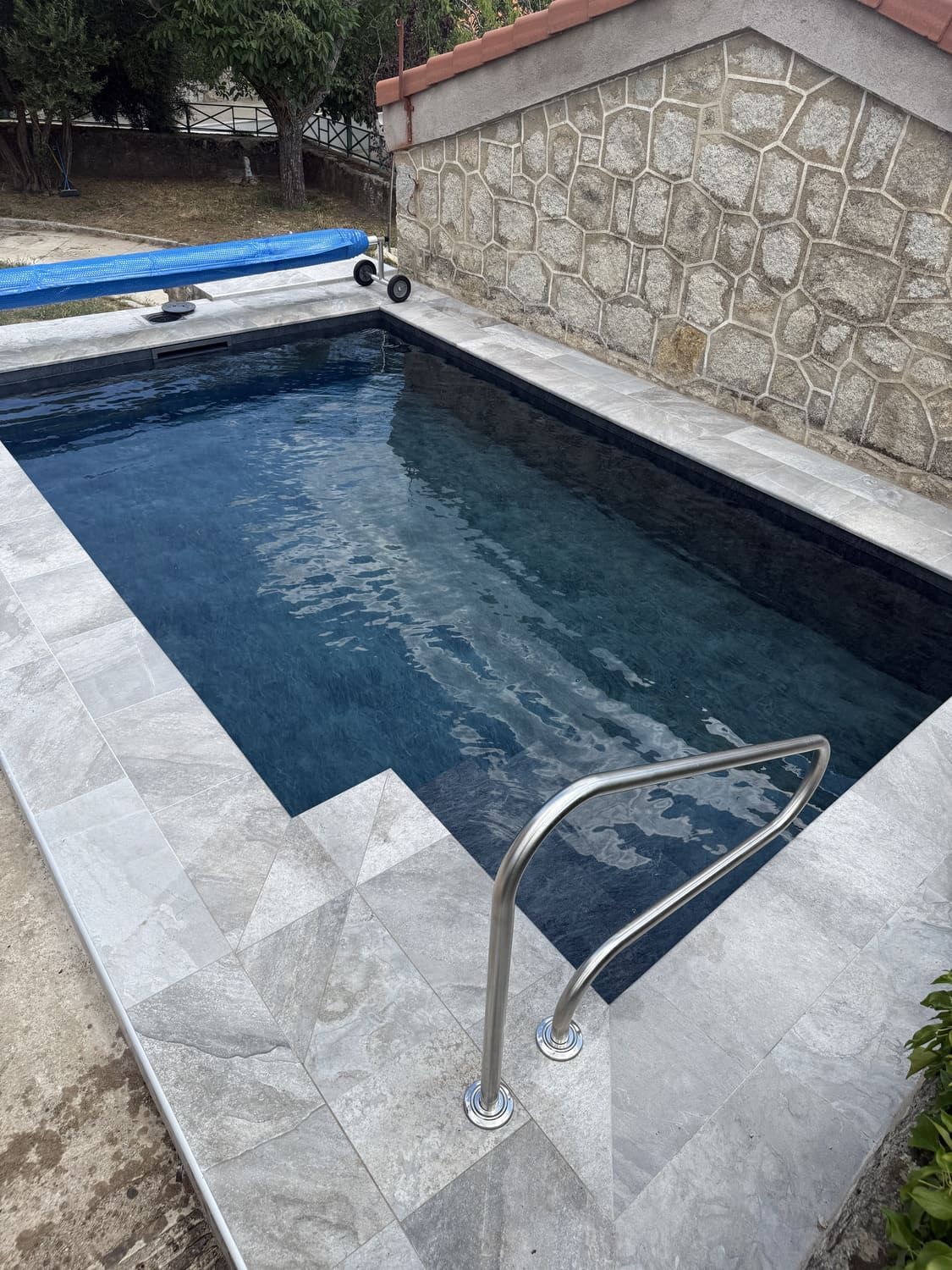 Piscina rectangular con escalones y zona circundante de baldosas grises, barandilla de acero inoxidable y fondo de pared de piedra.