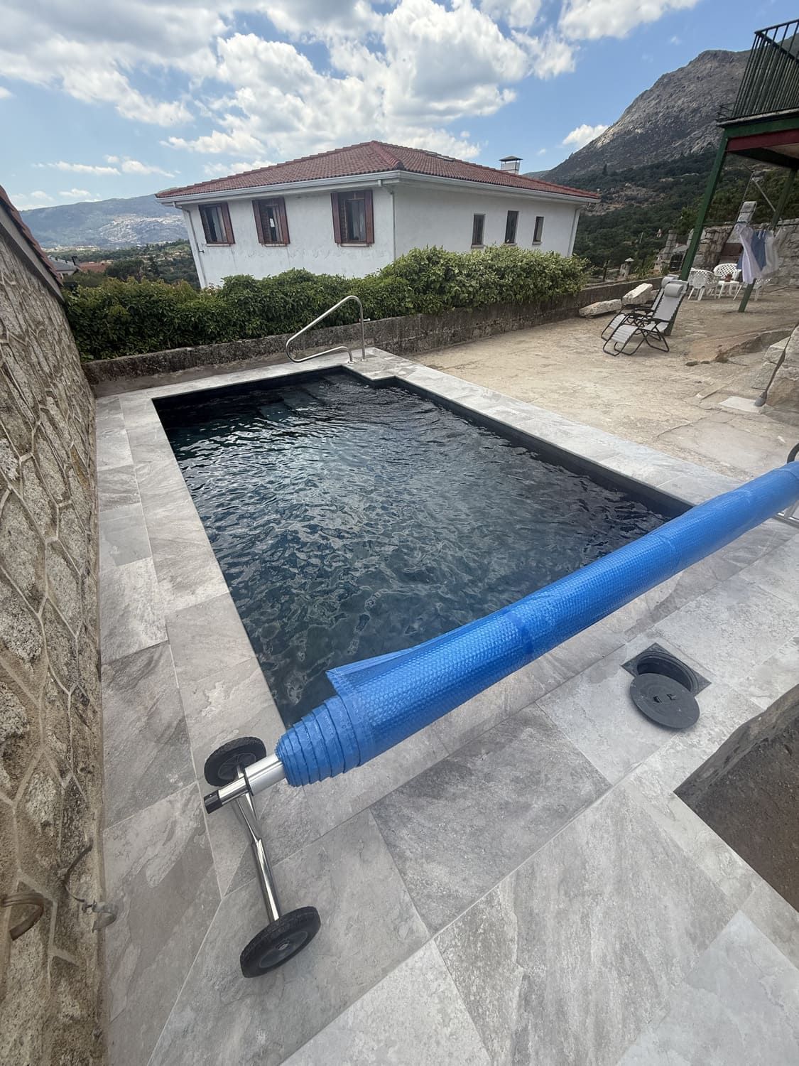 Piscina con cubierta azul, rodeada de patio de piedra, con una casa y montañas al fondo.