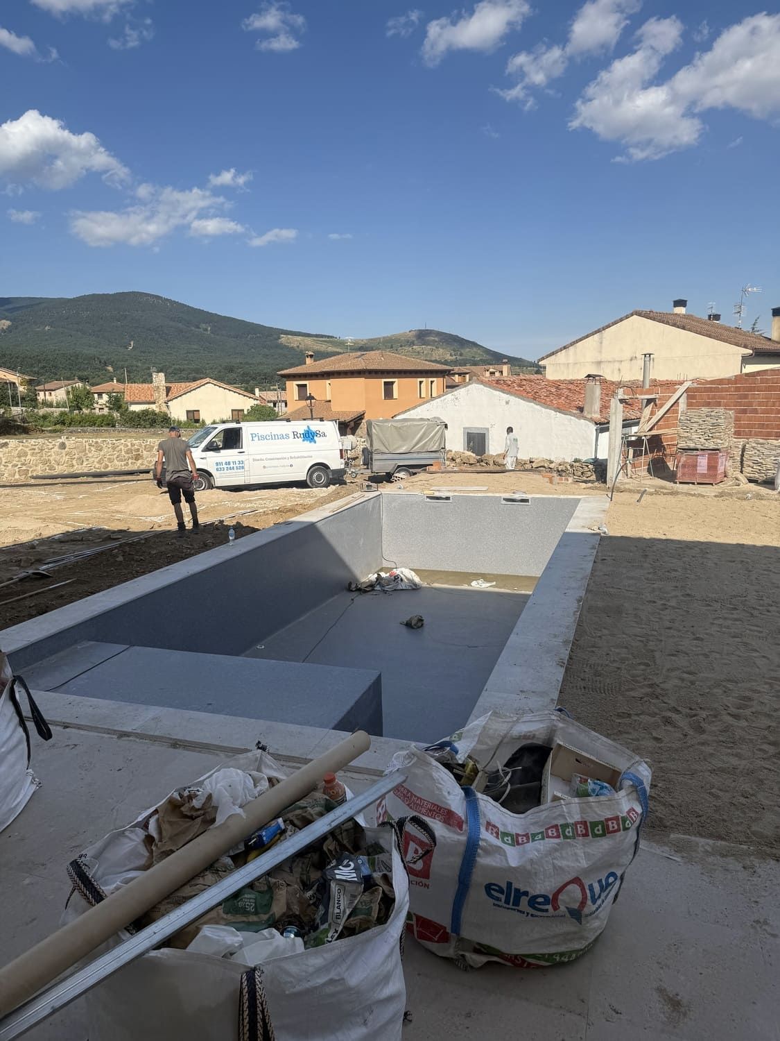 Una piscina parcialmente construida con un trabajador cerca, una camioneta blanca y casas bajo un cielo azul.