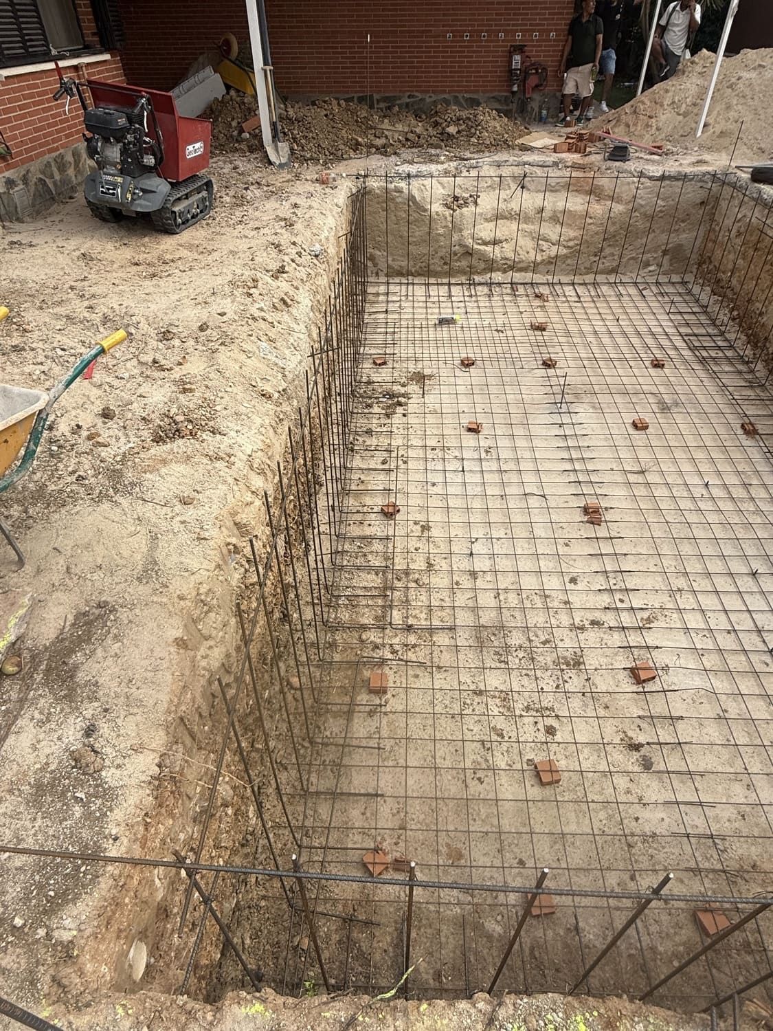 Sitio de excavación de piscina, que muestra una rejilla de barras de refuerzo en el suelo, soportes de ladrillo y una miniexcavadora.