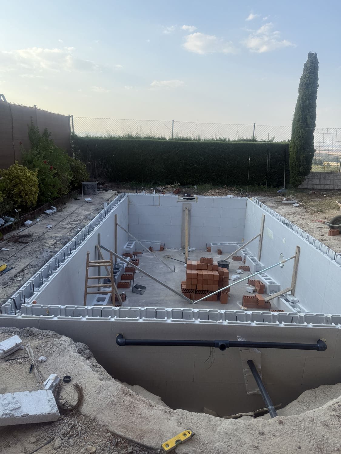 Piscina en construcción, forma rectangular, bloques grises, pila de ladrillos en el interior. Tuberías extendidas. Exterior, cielo nublado.