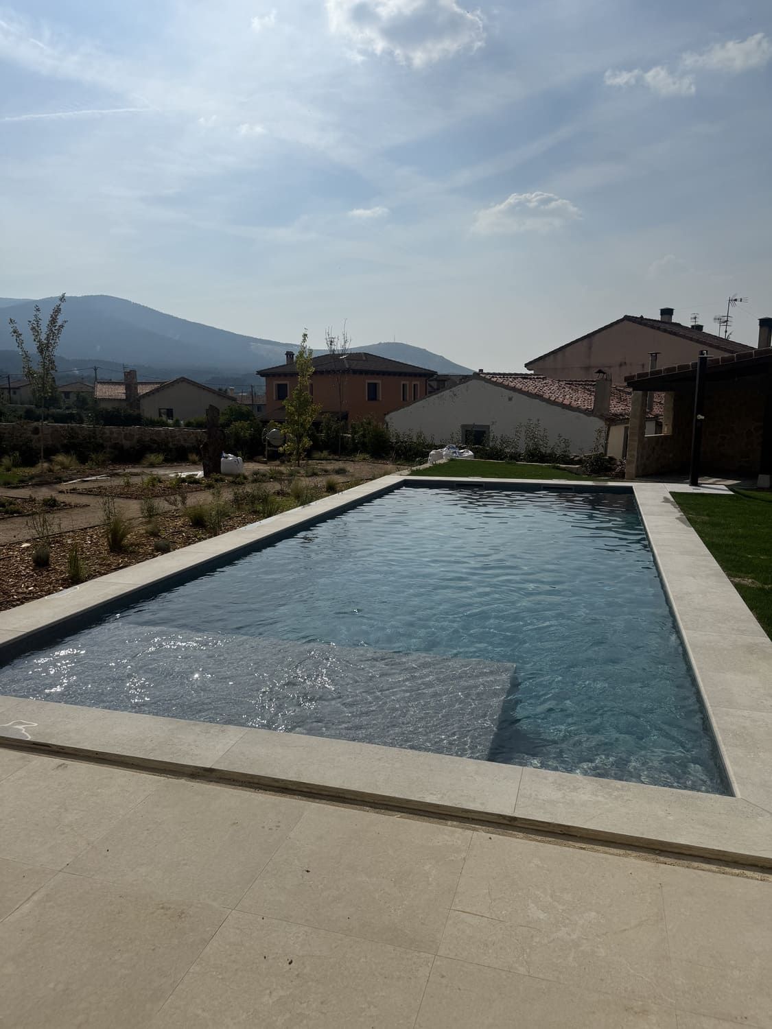 Piscina rectangular con marco de piedra clara; casas y montañas al fondo.