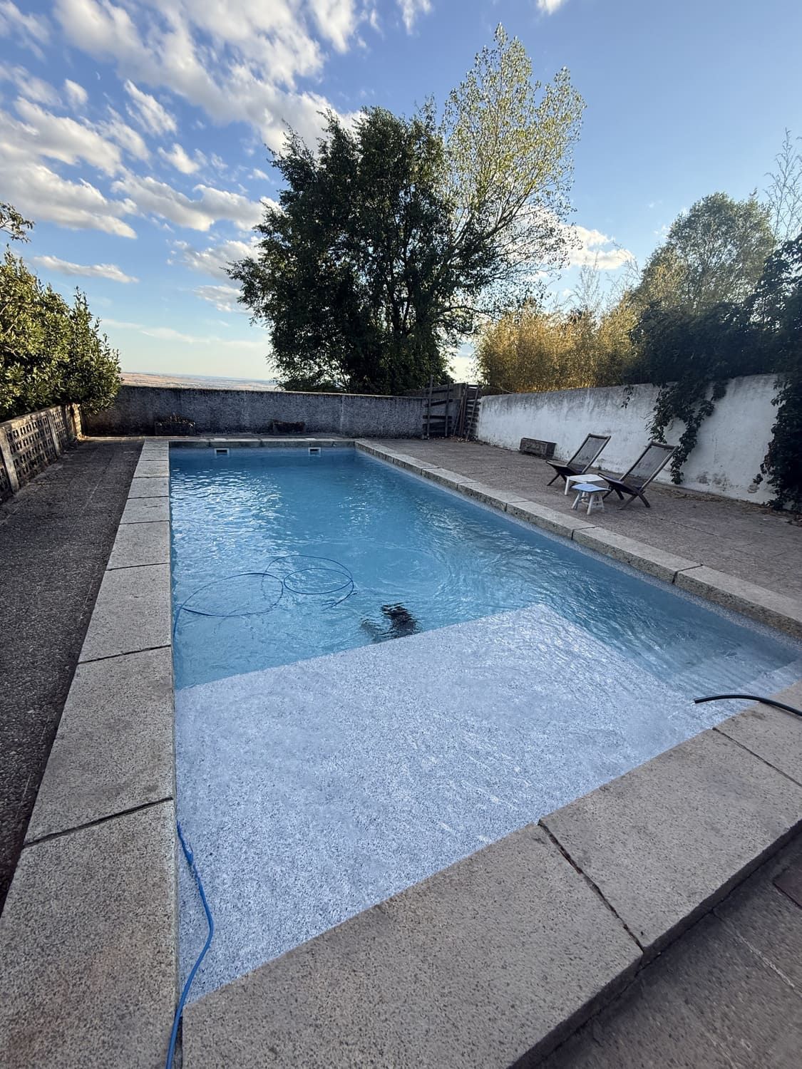 Piscina exterior rectangular con borde de piedra. Cielo azul nublado.