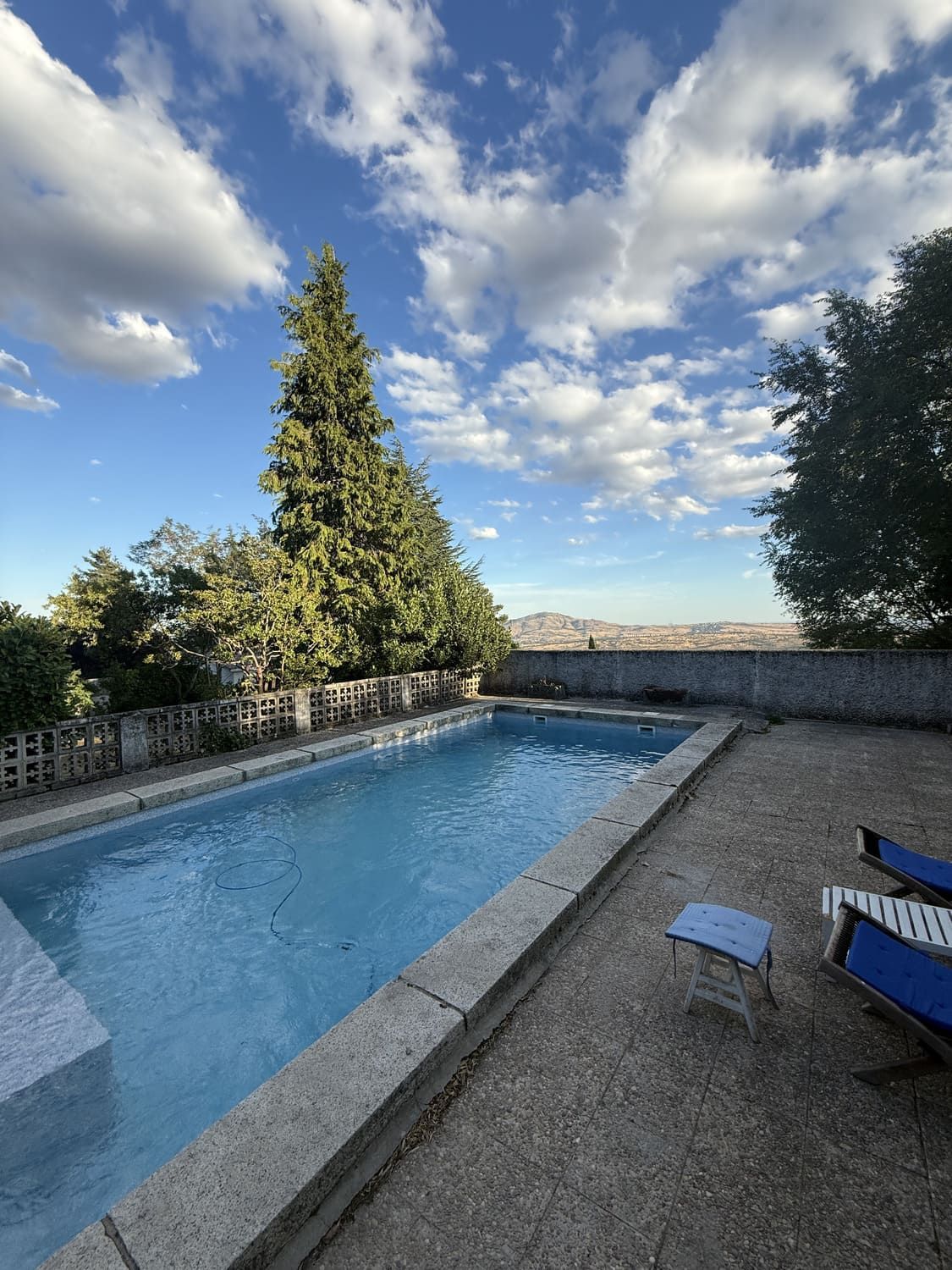 Piscina con agua azul cristalina, marco de piedra y vista lejana de la ciudad bajo un cielo nublado.