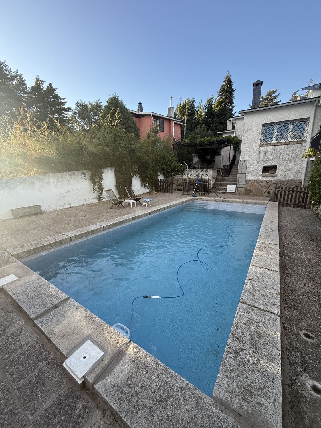 Una piscina exterior rectangular rodeada de piedra, con edificios y árboles al fondo.