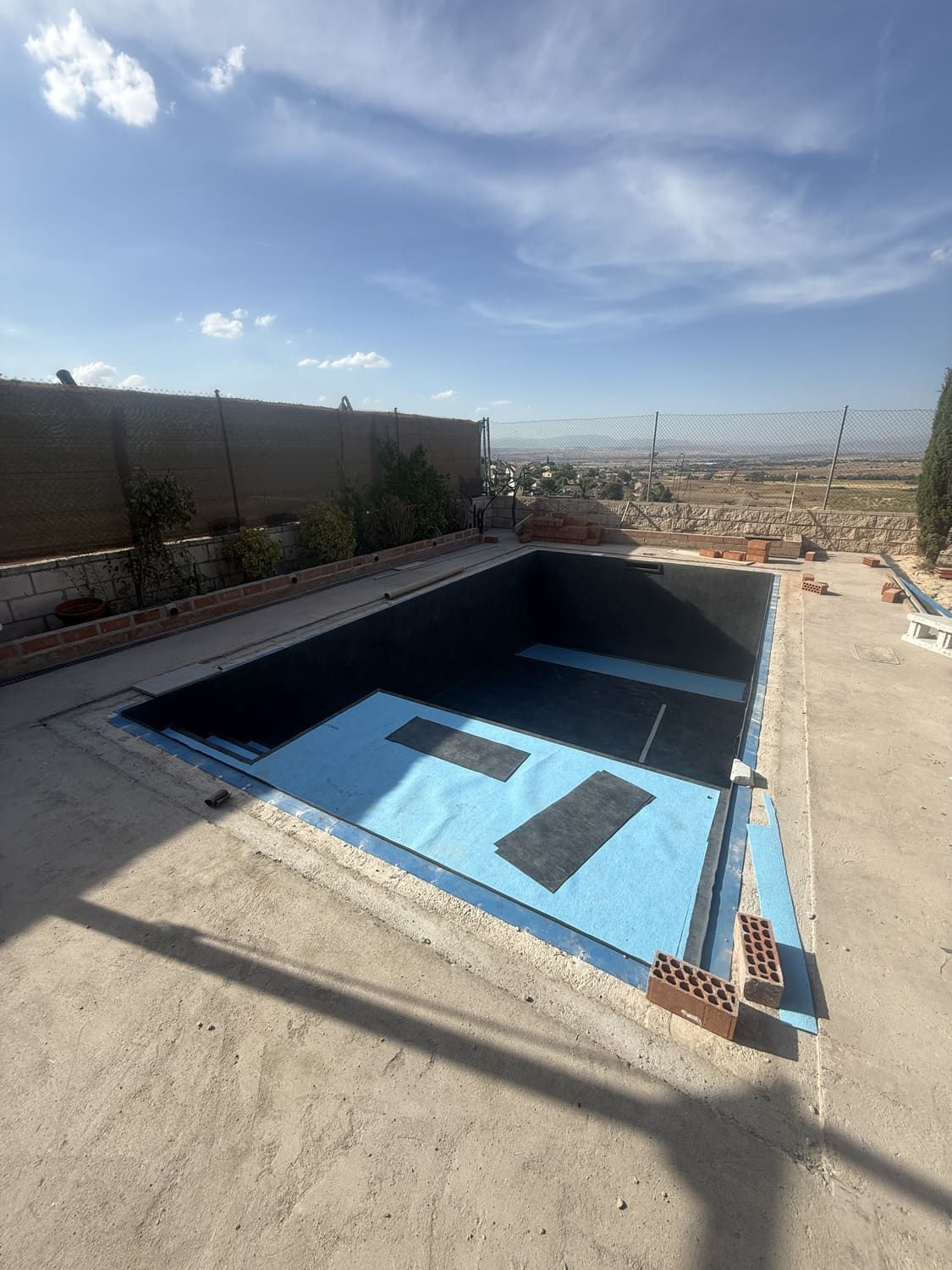 Piscina rectangular vacía en construcción, capas protectoras azules, revestimiento oscuro, entorno exterior soleado.