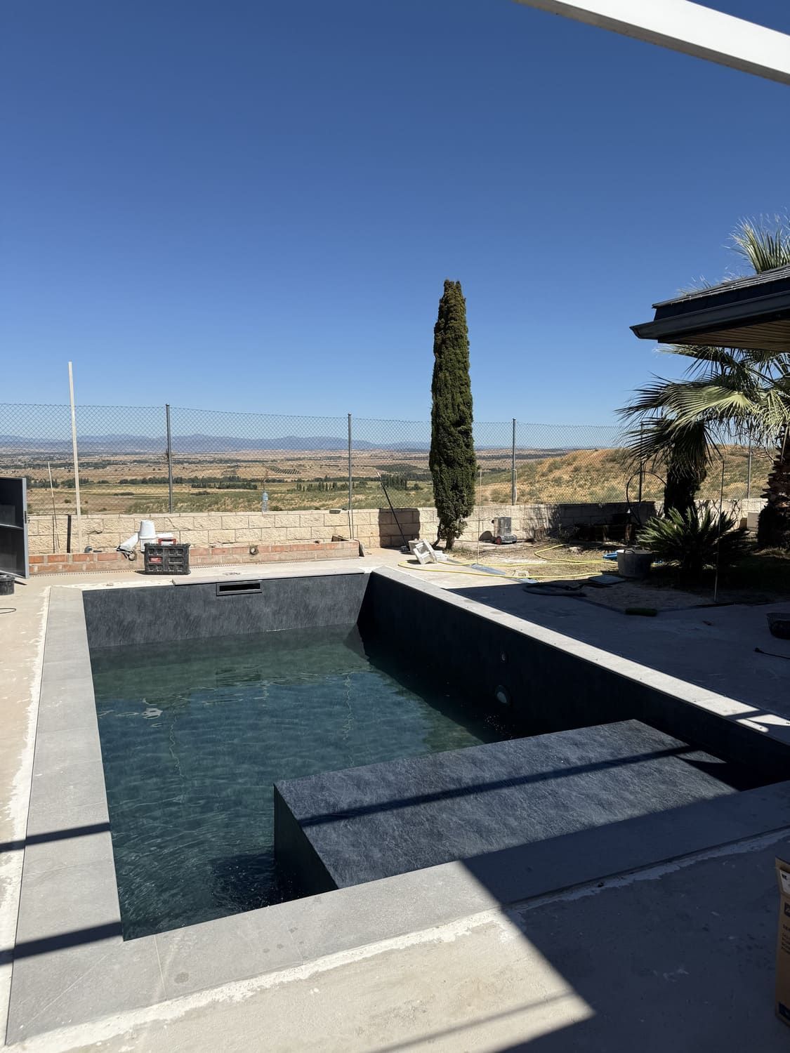 Piscina rectangular de azulejos oscuros con escalones; árbol alto y delgado; cielo azul claro; paisaje seco y montañoso en el fondo.