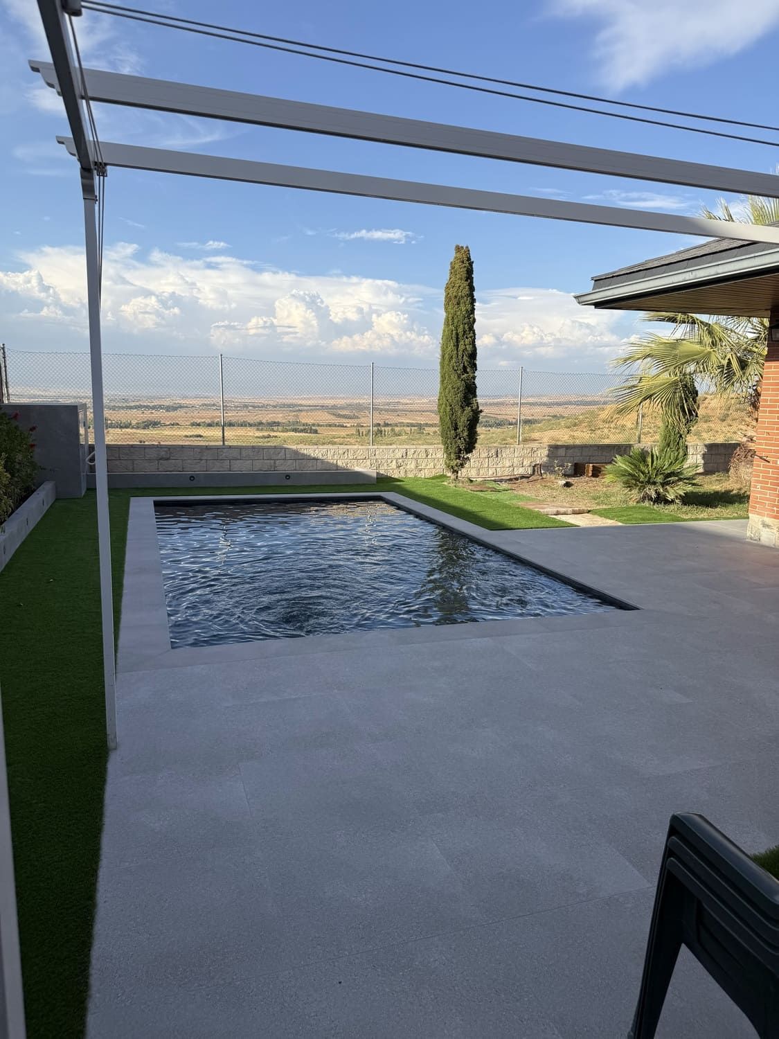 Piscina y patio con pérgola, con vista a un paisaje bajo un cielo azul.