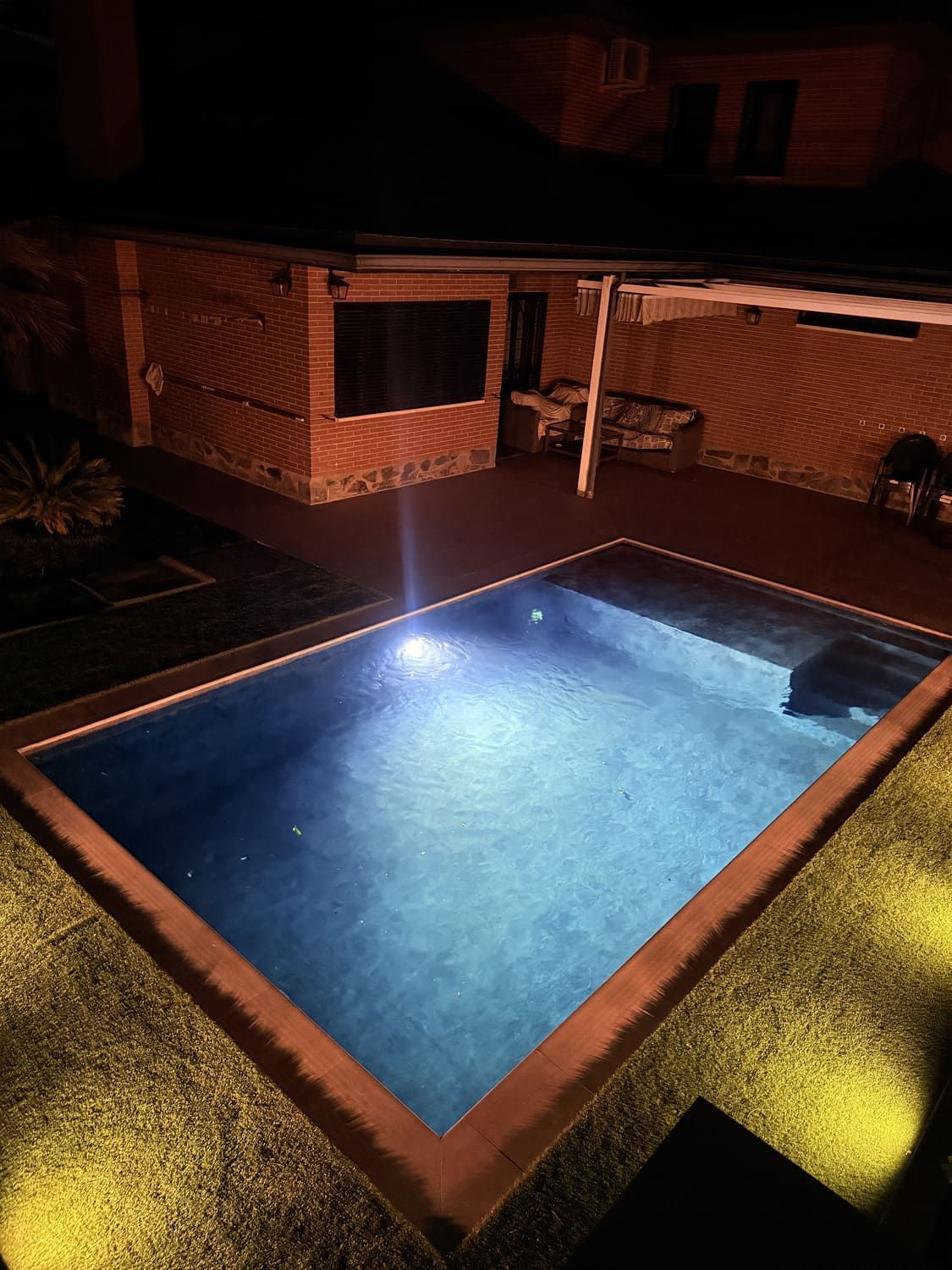 Vista nocturna de una piscina iluminada rodeada de césped artificial, paredes de ladrillo y una pequeña estructura.