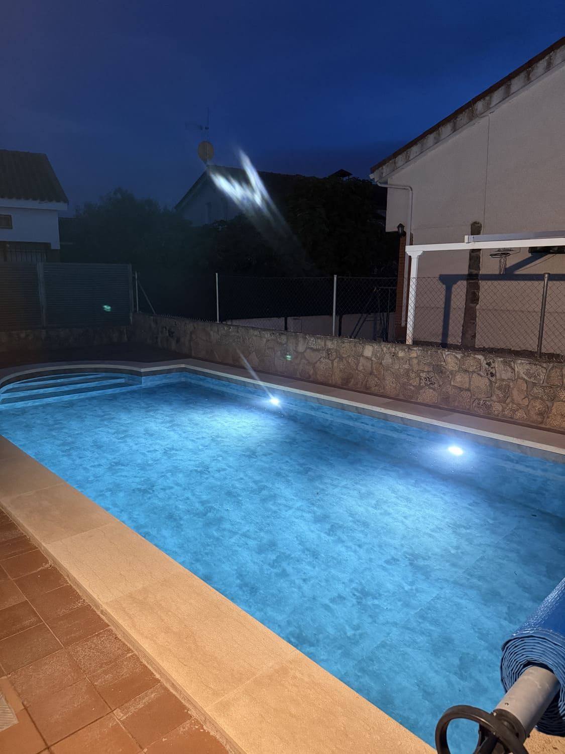 Piscina iluminada de noche con agua azul, marco de ladrillo y una casa de fondo.