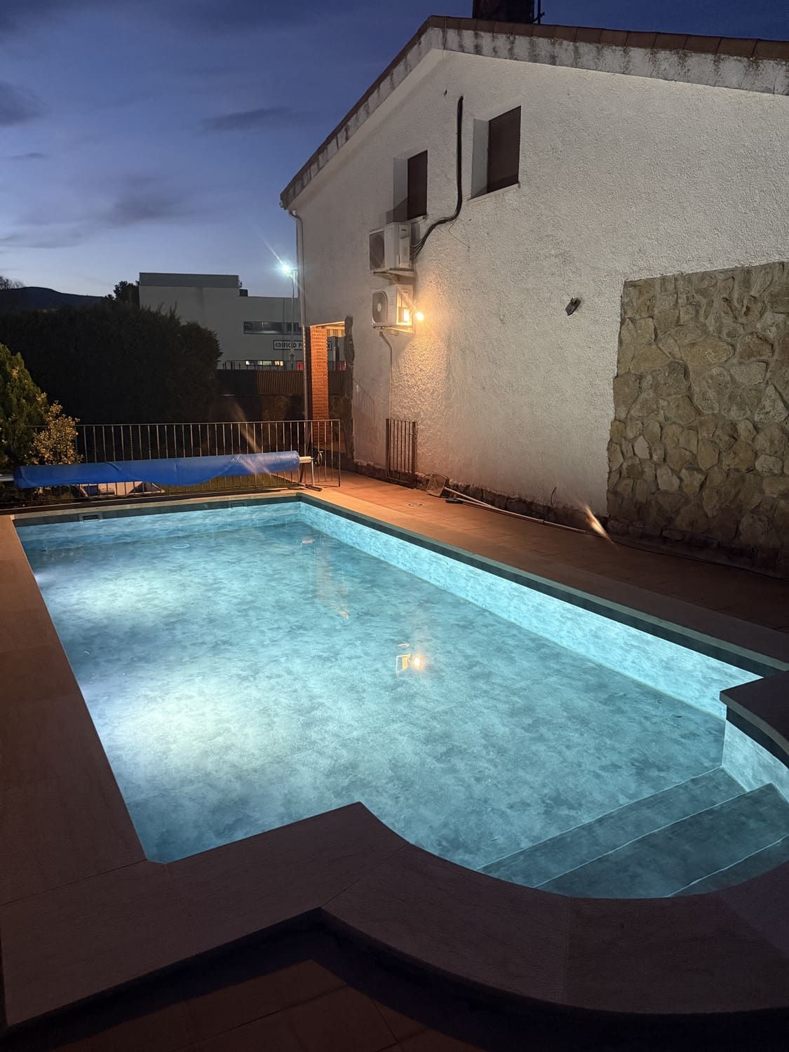 Vista nocturna de una piscina junto a una casa blanca con una sección de muro de piedra.