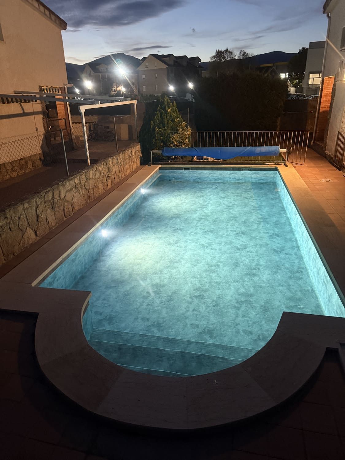Vista nocturna de una piscina iluminada con terraza circundante y edificios al fondo.