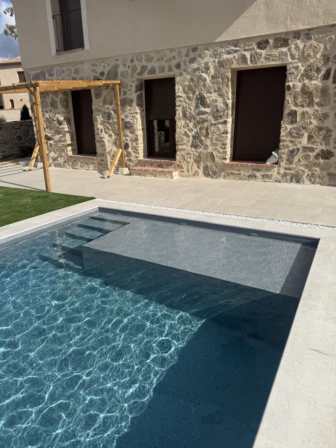 Piscina con escalones, junto a un edificio de piedra con persianas marrones. La piscina está rodeada de hormigón gris, con césped verde a un lado.
