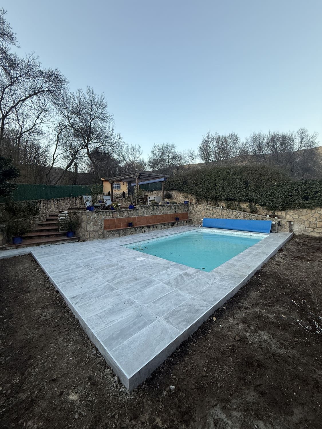 Piscina con cubierta azul, rodeada de patio gris, muro de piedra y árboles desnudos.