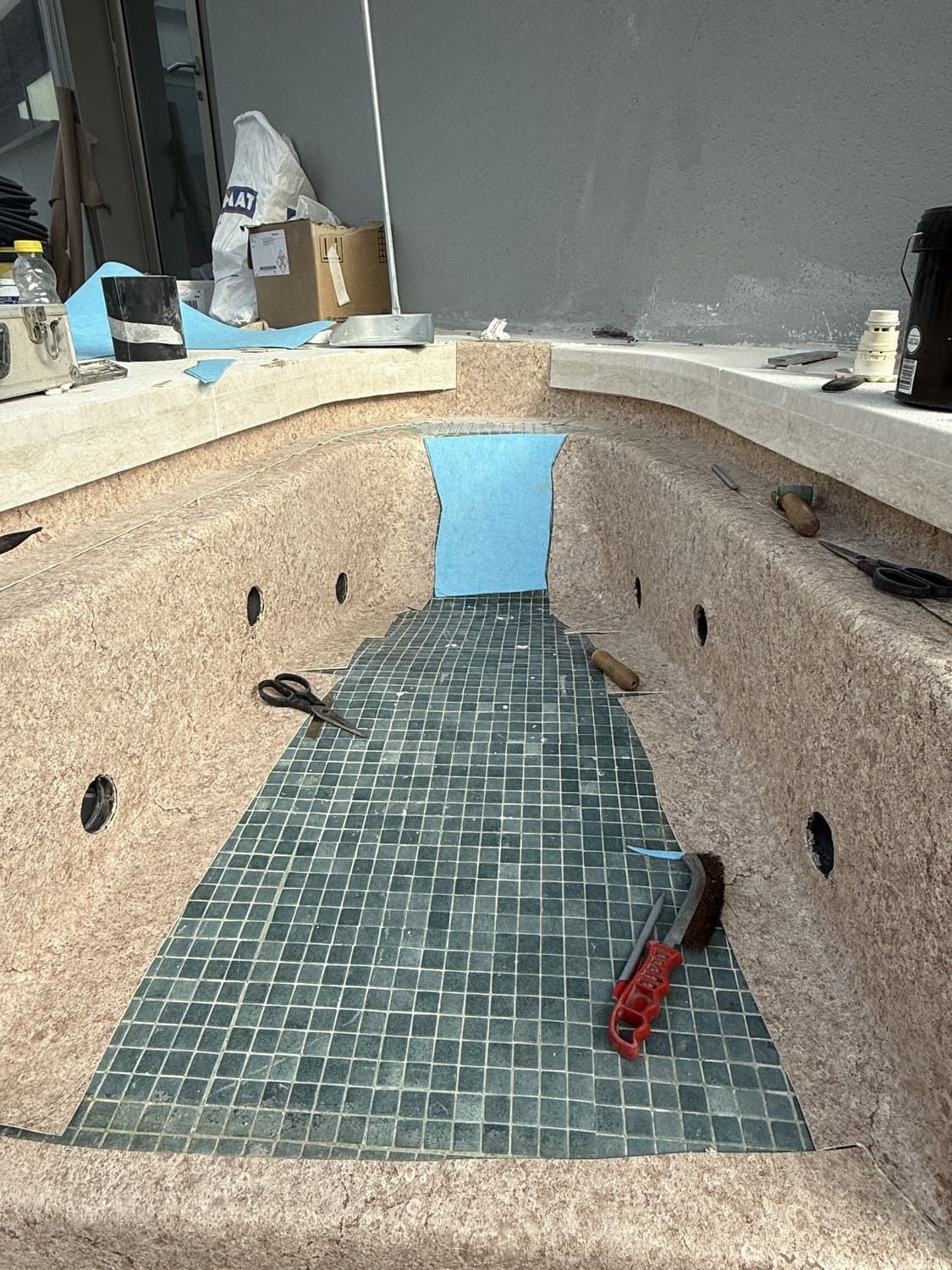 Una piscina parcialmente embaldosada con azulejos azules en proceso de instalación. Un cortador rojo yace sobre los azulejos.