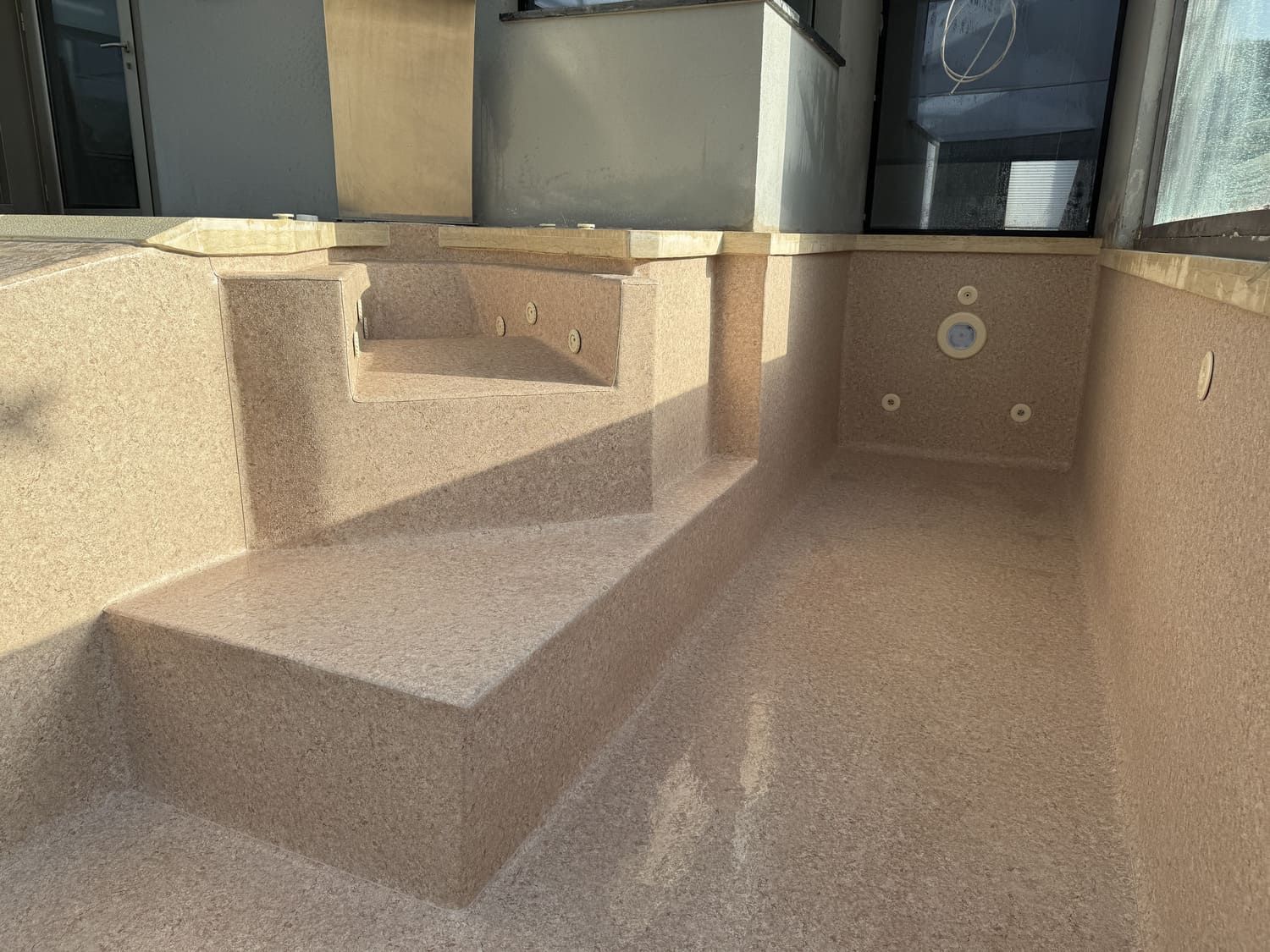 Piscina vacía de color marrón claro moteada, con escalones, chorros y entradas de agua, frente a un edificio.