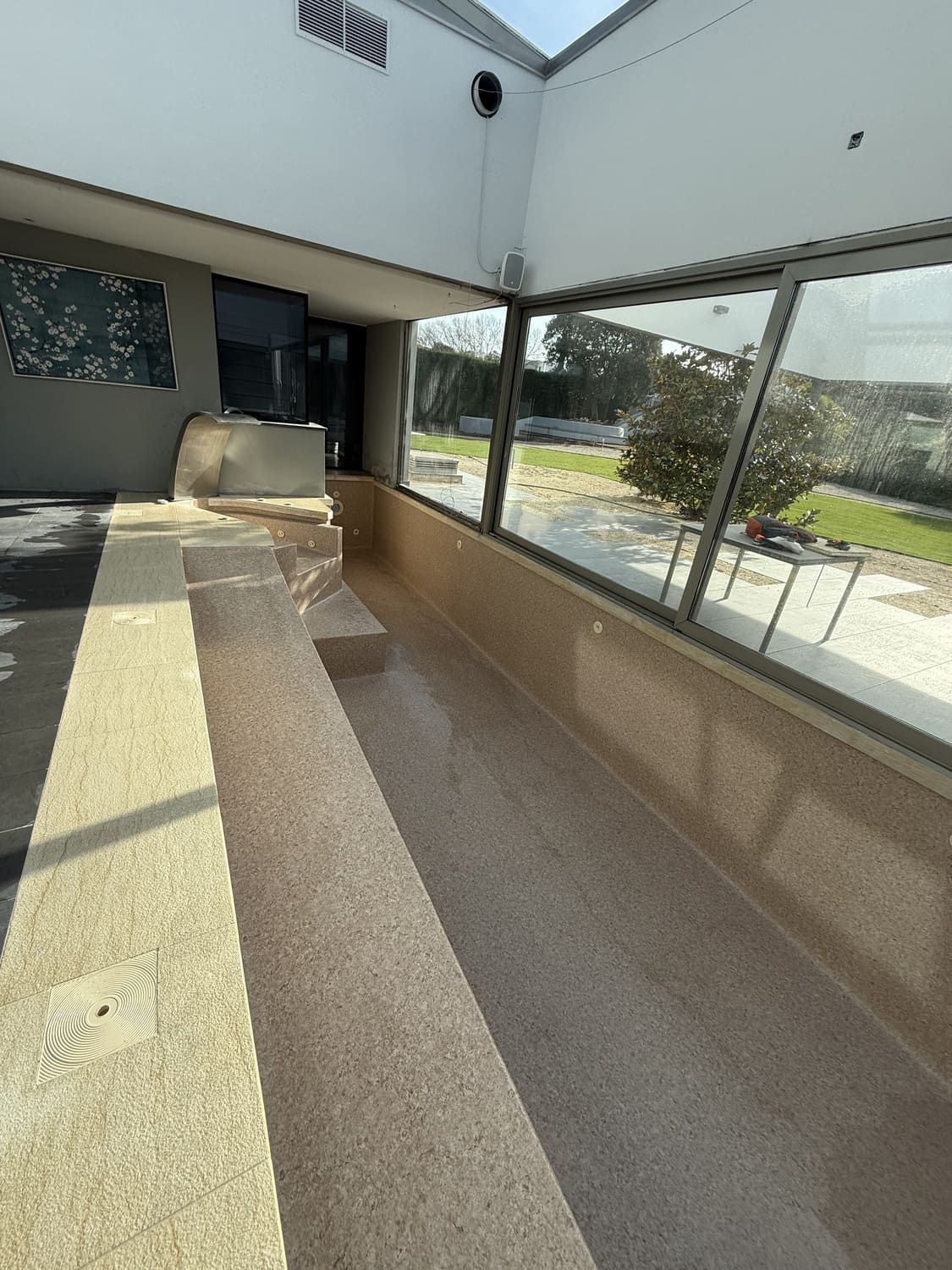 Piscina larga y estrecha, de azulejos de color beige, junto a una pared de cristal, bajo el alero blanco de un edificio.