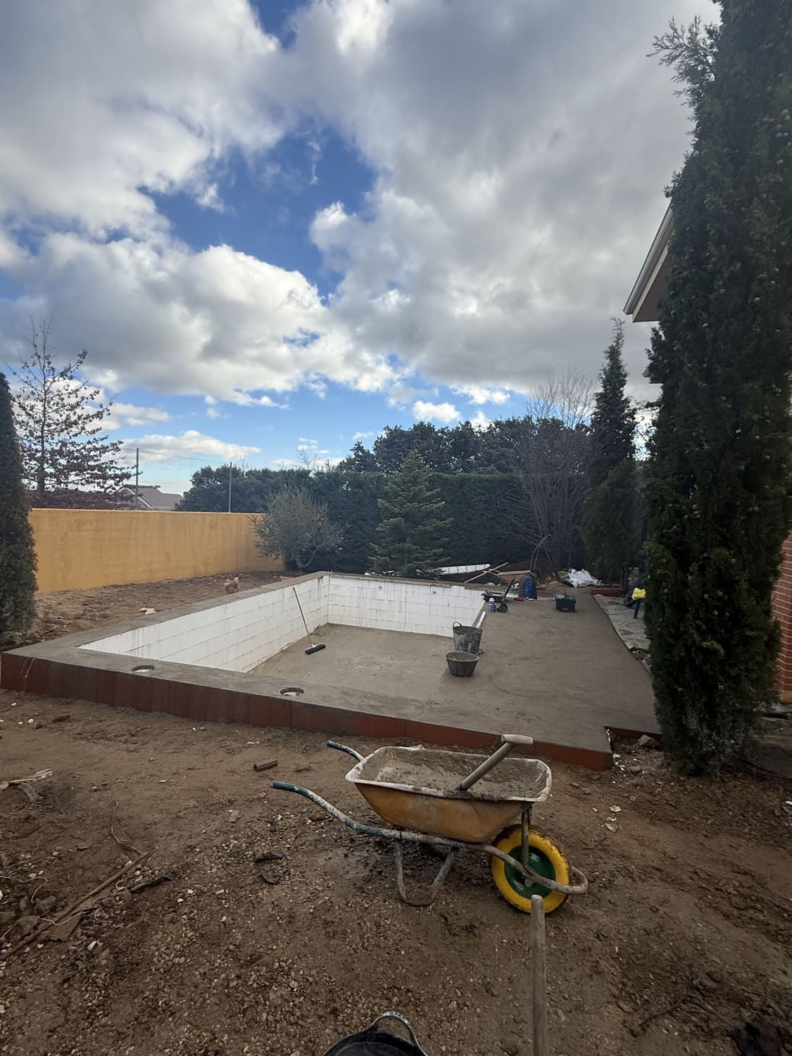 Piscina vacía en construcción; carretilla amarilla en primer plano, cielo nublado.