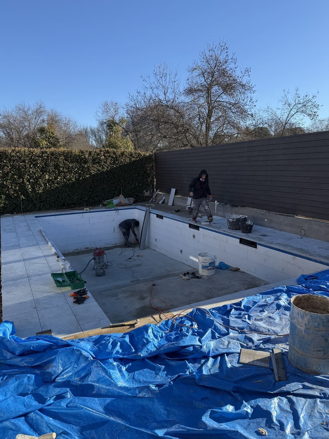 Piscina en construcción en un patio. Se ven trabajadores, herramientas, lona azul, hormigón y vallas.