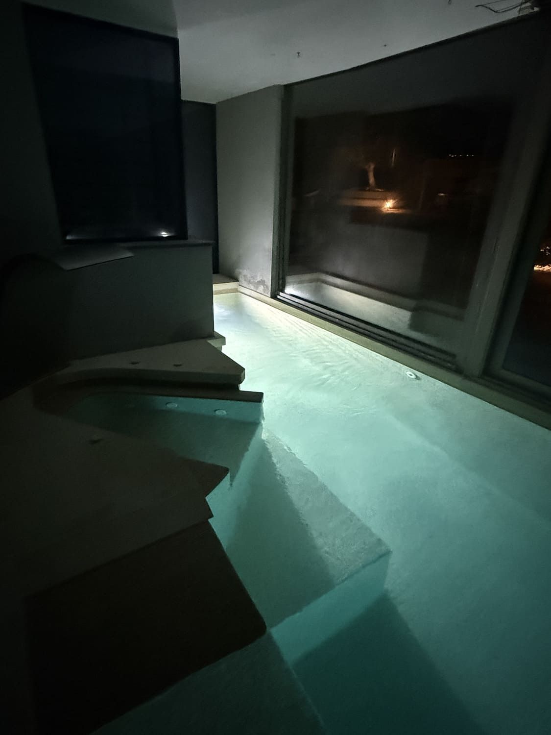 Piscina cubierta con escaleras incorporadas, iluminada por agua turquesa y luz exterior a través de un gran ventanal.