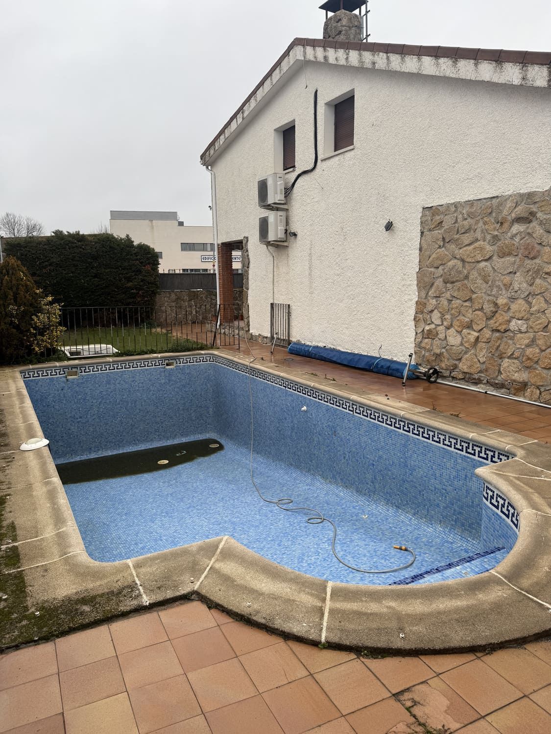 Piscina vacía de azulejos azules junto a una casa blanca con muro de piedra. Día nublado.