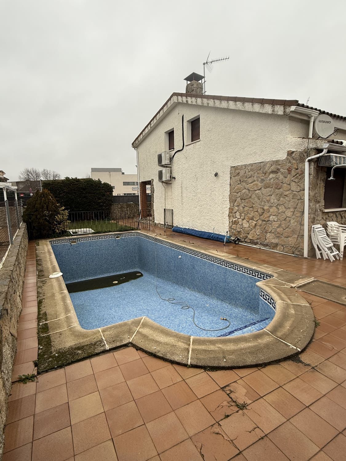 Patio trasero con una piscina vacía de azulejos azules, junto a una casa blanca de dos pisos con un muro de piedra. Cielo nublado.