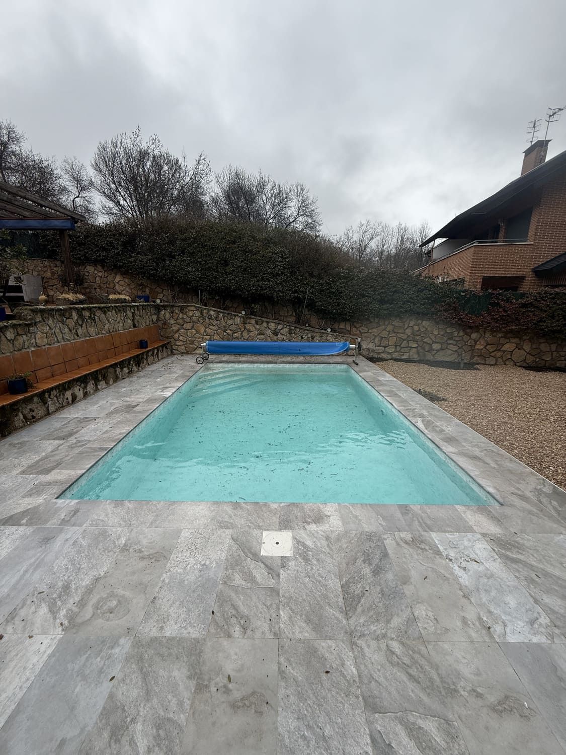 Piscina rectangular con cubierta azul, rodeada de baldosas de piedra gris. Cielo nublado, árboles y una casa al fondo.