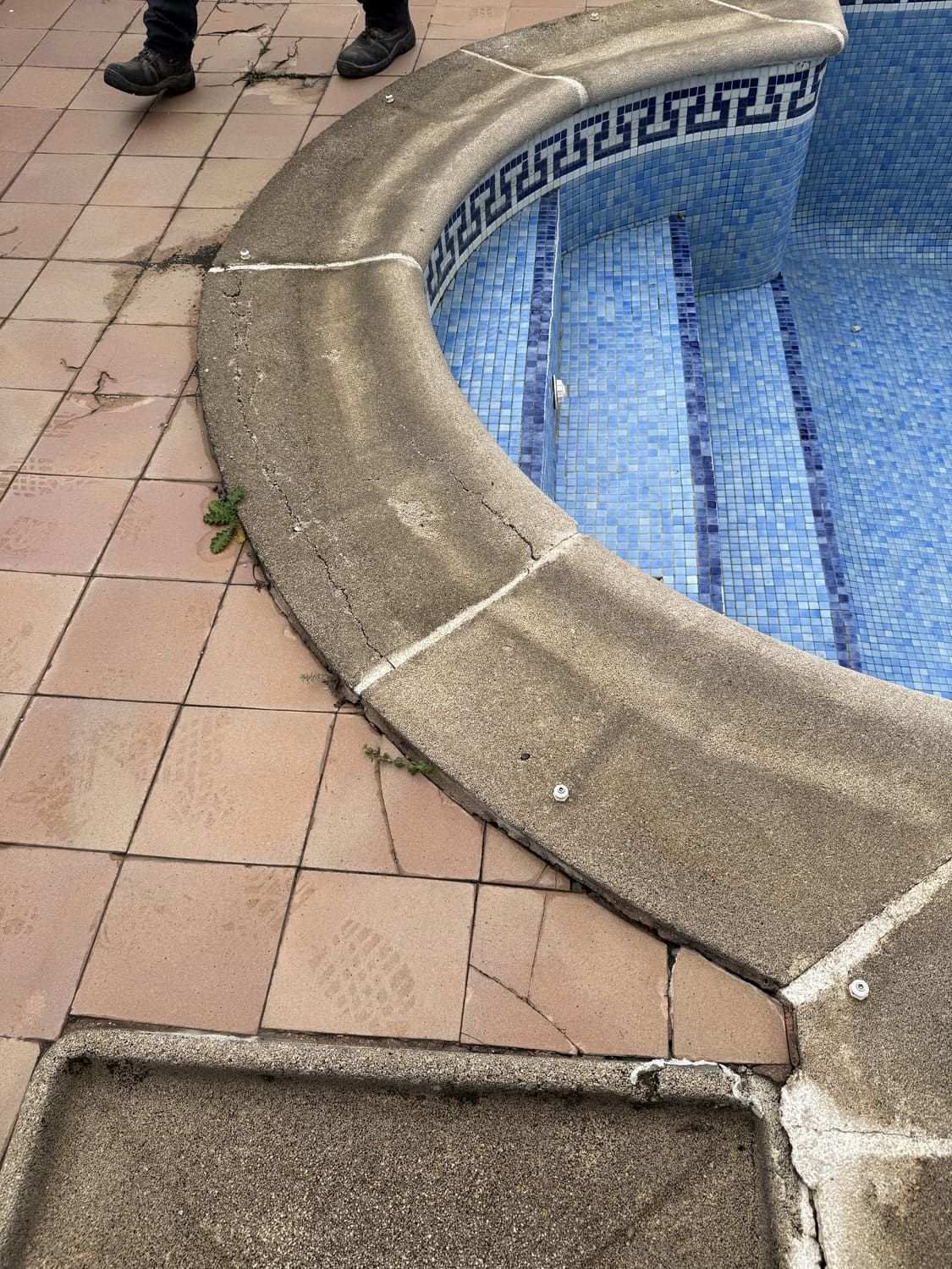 Junto a la piscina con azulejos azules y borde curvo de hormigón. Se ven las piernas de alguien cerca del borde sobre azulejos rojos.