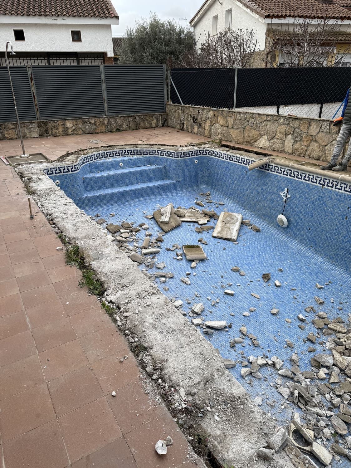 Piscina en proceso de renovación con hormigón visto y baldosas rotas, rodeada de patio de ladrillo.