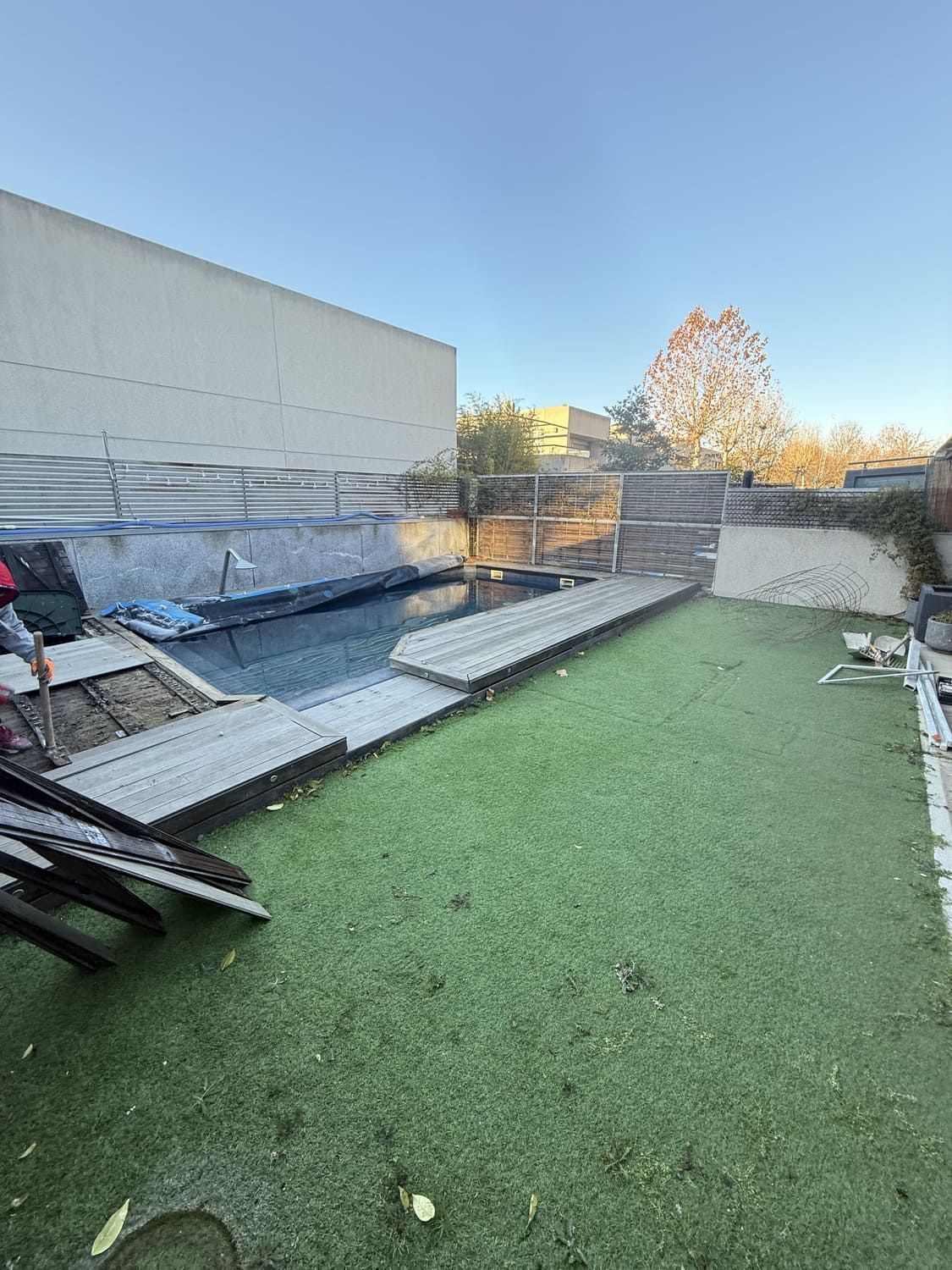 Zona de piscina con piscina parcialmente cubierta, terraza de madera y césped. Un edificio al fondo.