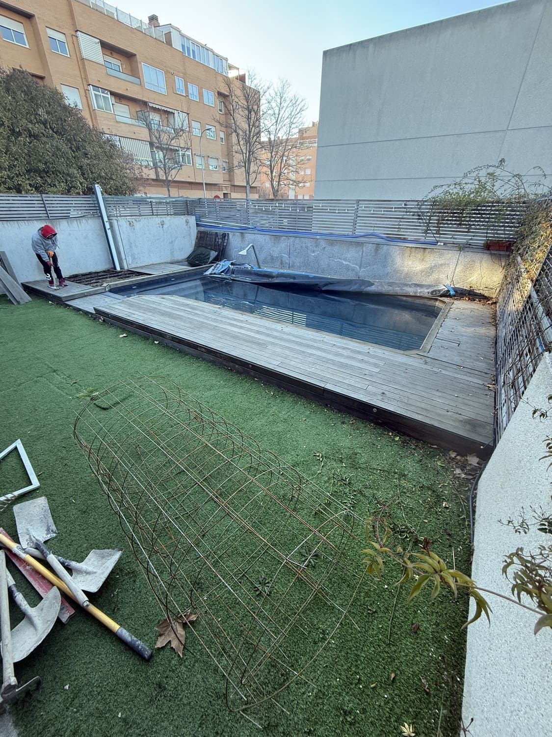 Piscina en azotea rodeada de césped artificial, una persona trabaja cerca, entorno urbano.