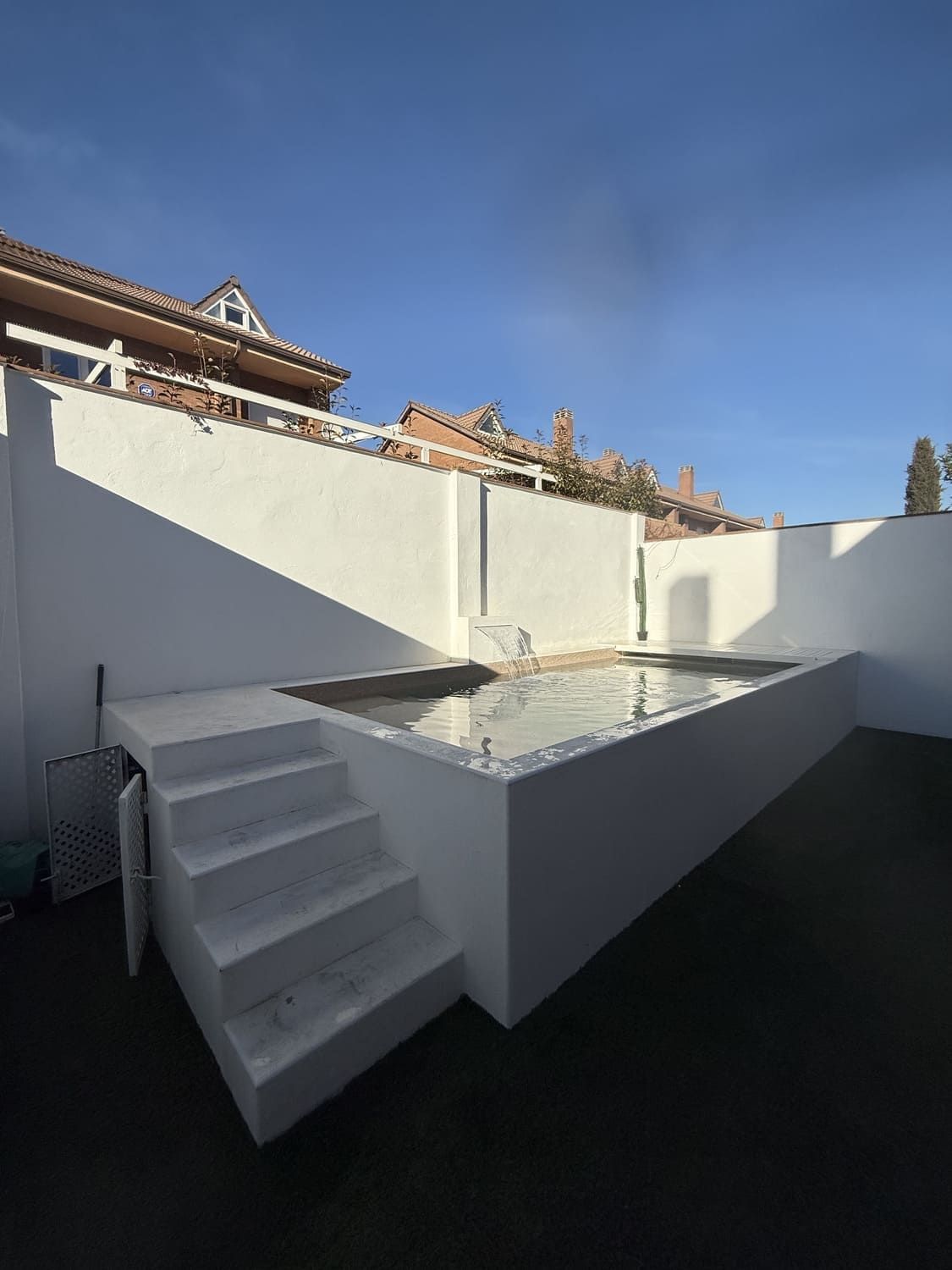 Piscina rectangular blanca con escaleras, frente a paredes blancas bajo un cielo azul.