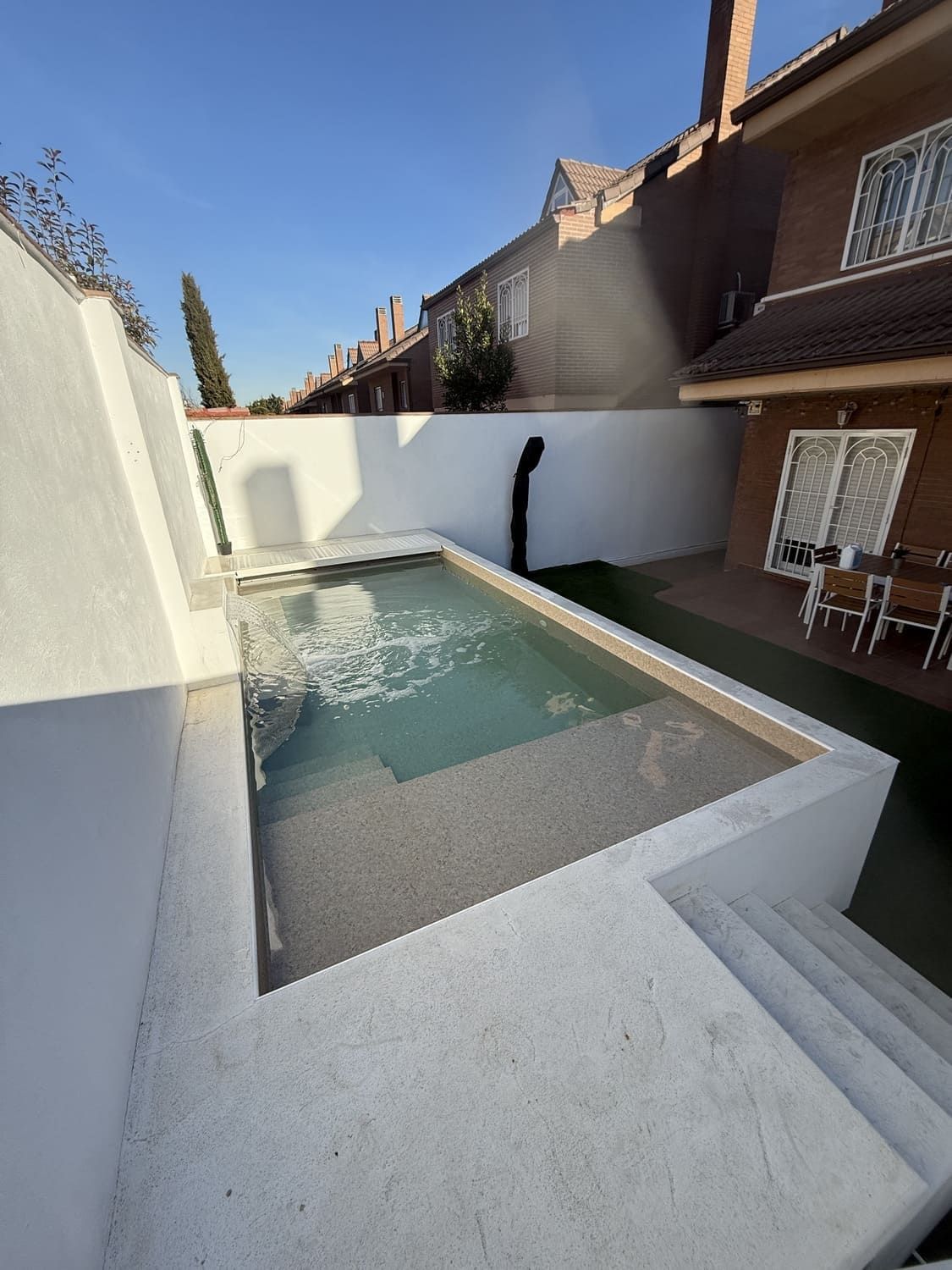 Pequeña piscina rectangular rodeada de paredes blancas y escaleras, al lado de una casa con techo de tejas.